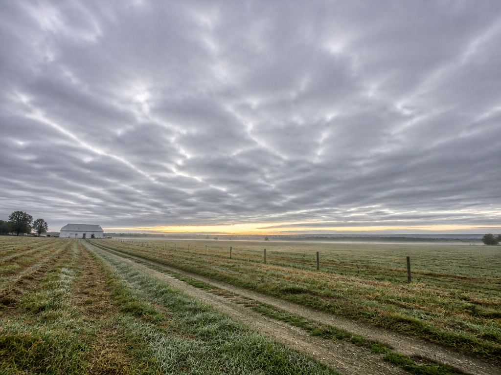 A photorealistic dawn landscape photograph of an open Pennsylvania farm field under a vast…