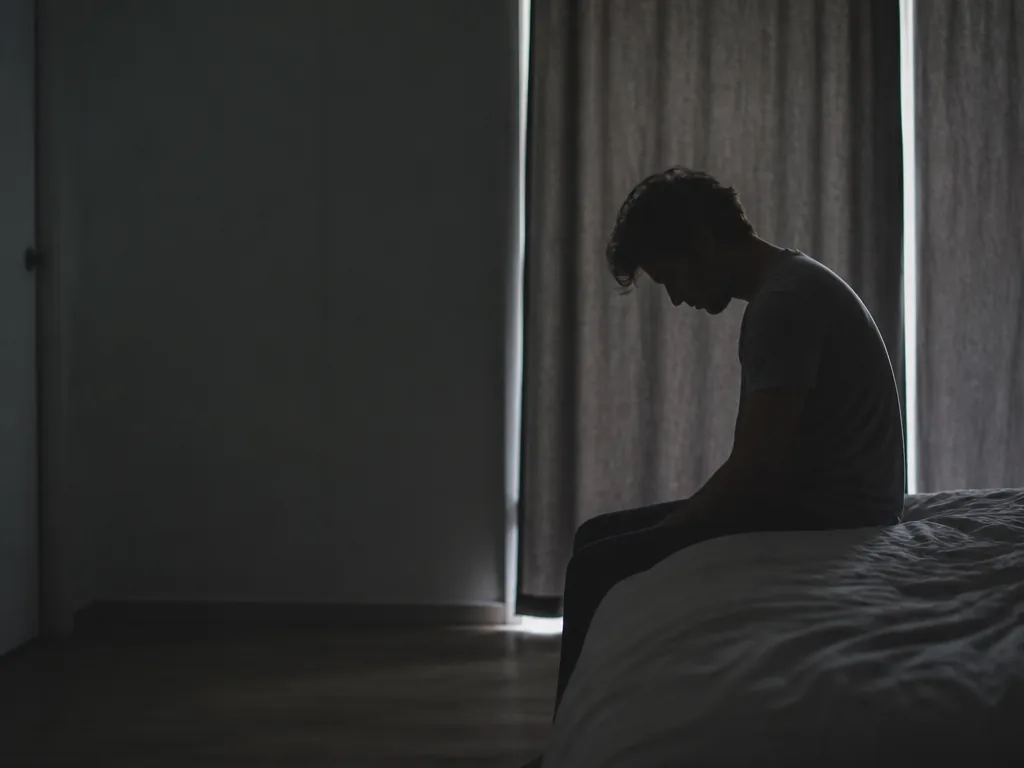 A person seated on the edge of a bed at first light in a…