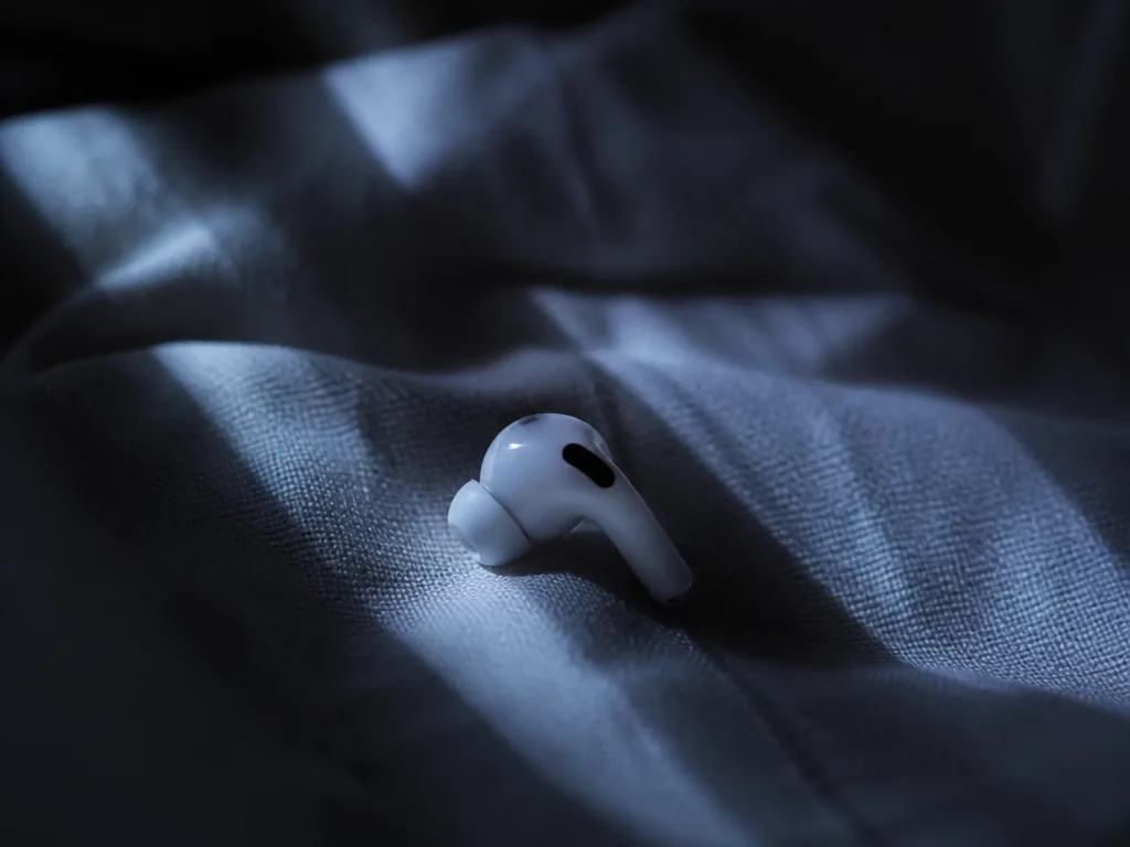 A photorealistic overhead close-up of a single white wireless earbud resting on rumpled pale…